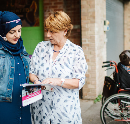 Image shows two ladies talking together while looking at a leaflet. In the background is a person in a wheelchair.
