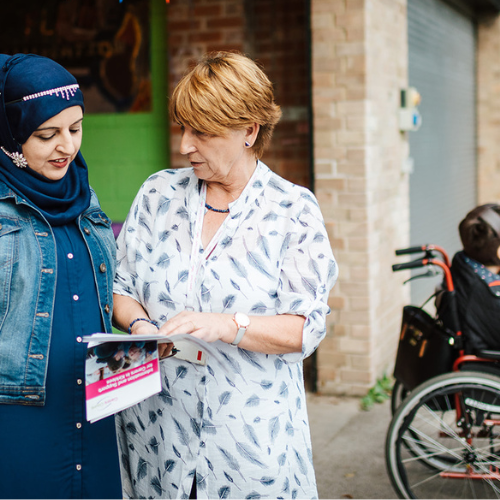 Image shows two ladies talking together while looking at a leaflet. In the background is a person in a wheelchair.